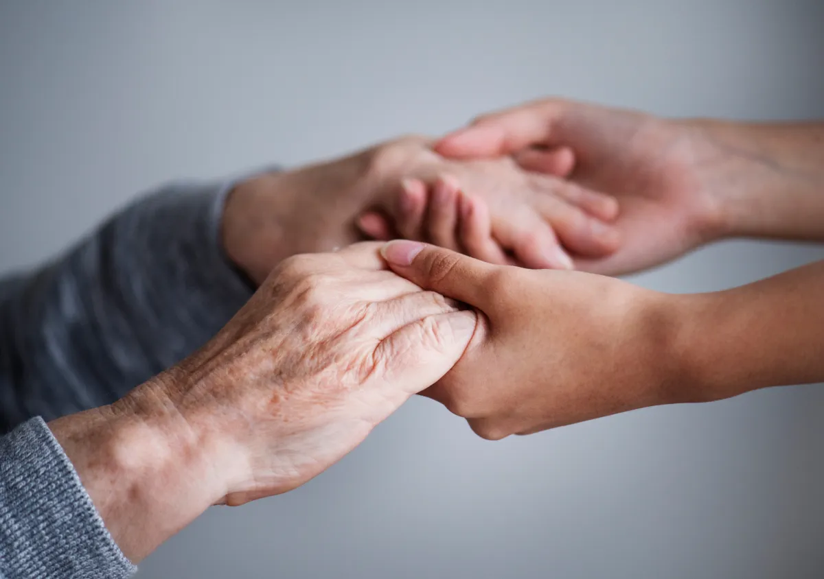 Madre anziana e figlia che si prendono per mano, a simboleggiare l’affetto e la cura che la figlia ha per la madre con difficoltà di autosufficenza.